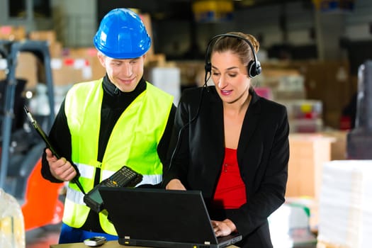 Teamwork - warehouse clerk or warehouse worker and customer service representative using laptop and headset in a warehouse of a freight forwarding company,she points at the camera
