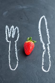 One fresh whole strawberry and chalk drawings of knife and fork on dark background