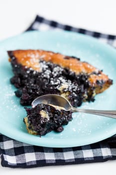 Closeup of slice of delicious blueberry pie on plate with spoon resting on checkered napkin over light background
