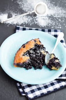 Closeup of slice of delicious blueberry pie on plate with spoon resting on checkered napkin with icing sugar sprinkled in background