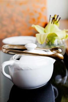 Vertical Interior corner detail of black top table with tea and coffee utensils of sugar bowls and bud vase with a lily by a window