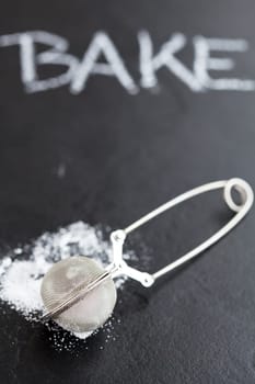Closeup of sieve and icing sugar with word bake written in chalk on dark textured background