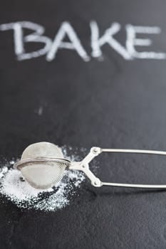 Closeup of sieve and icing sugar with word bake written in chalk on dark textured background