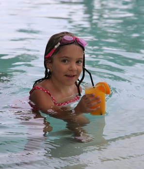 Beautiful happy little girl relaxing in the pool with some orange juice
