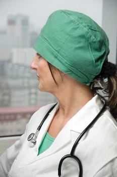 Side head and shoulders shot of a middle-aged doctor in a white coat,green hair covering and stethoscope placed on her neck looking thoughtfully doubtfully out of the window against an urban background