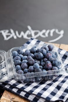 Fresh blueberries in plastic container on checkered napkin with word blueberry written in chalk on dark surface