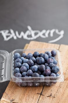 Fresh blueberries in plastic container on wooden serving surface with word blueberry written in chalk on dark surface