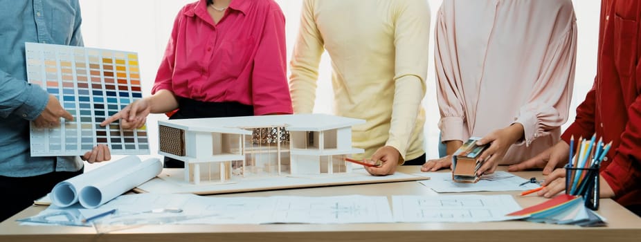 Cropped image of professional architect team brainstorm about house color selection while architect writes on blueprint document at table placed with blueprint and house model. Variegated.