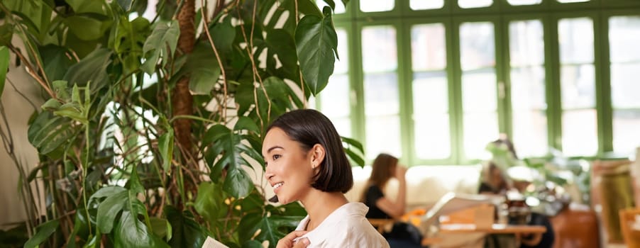 Vertical shot of asian brunette girl sitting elegant with book, reading in cafe and smiling, drinking coffee.