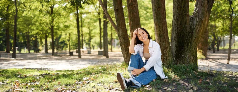 Young people. Beautiful asian girl sits near tree in park and rests, smiling and looking into distance, relaxing outdoors on fresh air in summer.