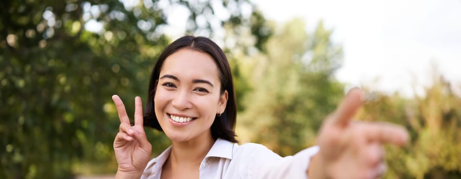 People and lifestyle. Happy asian woman takes selfie in park, photo on smartphone, smiling and looking joyful.