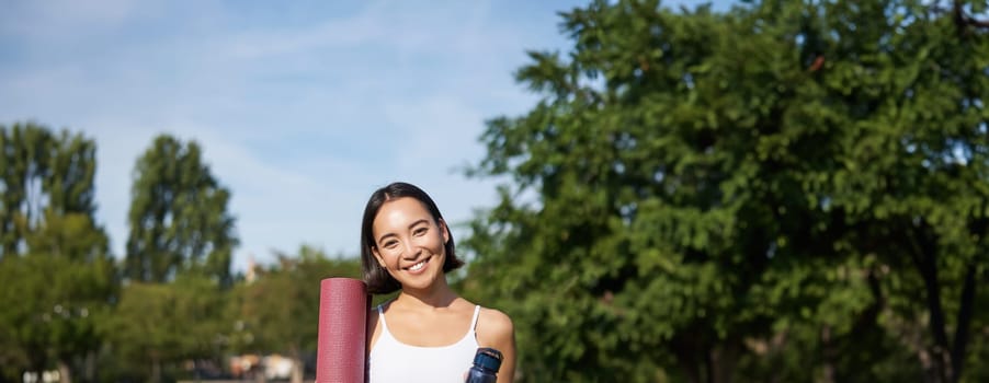 Portrait of young slim and healthy korean girl doing workout in park, standing with water bottle and rubber mat for execises on green lawn, smiling happily.