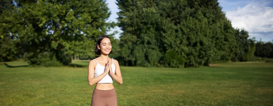 Vertical shot of smiling asian girl doing yoga, stretching exercises in park, standing on rubber mat on fresh air.