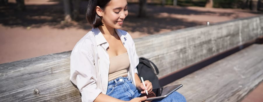 Young asian creative girl drawing with pen on graphic tablet, sitting outdoors in park, painting smth.