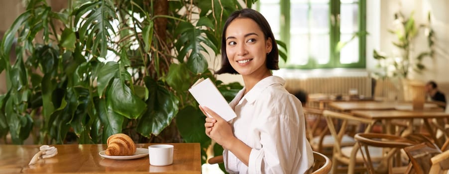 Tender, beautiful asian girl sitting with a book in cafe, reading and drinking coffee. People and lifestyle concept.