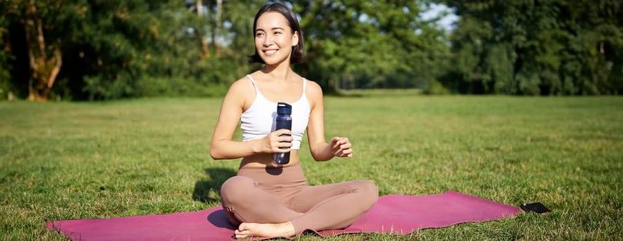 Smiling fitness girl sits on mat and drinks water after workout, finish yoga training in park, exercise on fresh air.