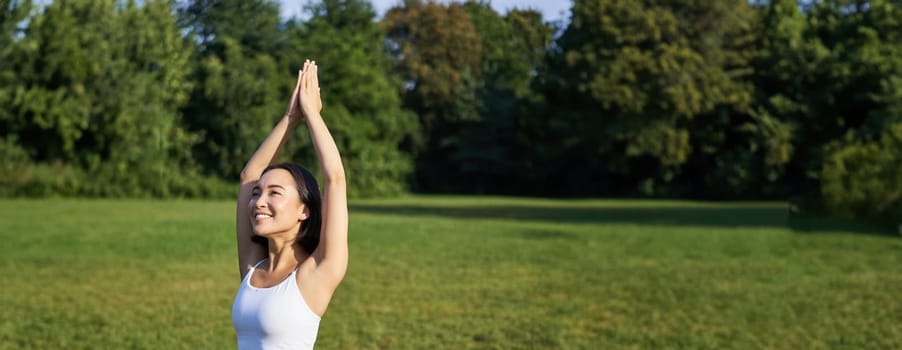 Vertical shot of young asian fitness girl doing yoga asana, standing in tree pose and smiling, execising in park on rubber mat.