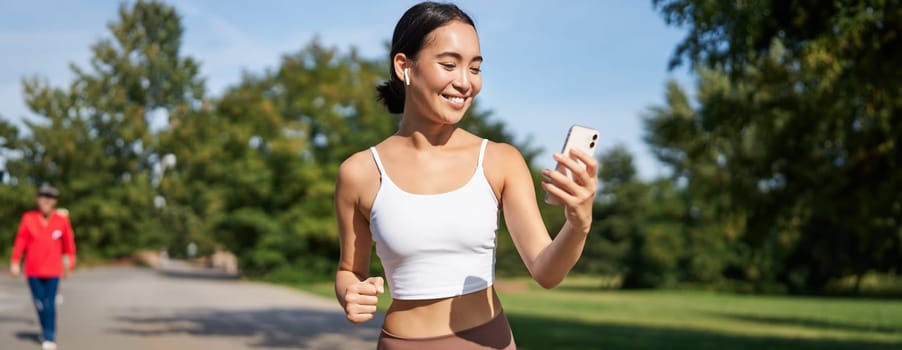 Fitness woman with water bottle and smartphone, jogging in park and smiling, looking at her mobile phone app, checking sport application.