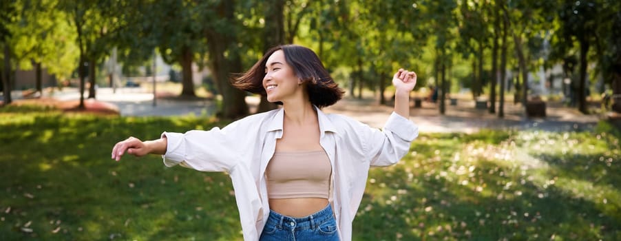 Freedom and people concept. Happy young asian woman dancing in park around trees, smiling and enjoying herself.