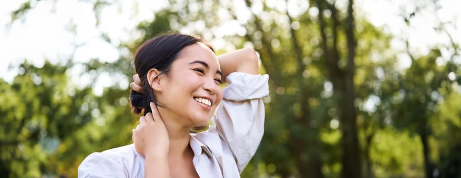 Beautiful young woman tying her hair while walking in park, smiling romantic, enjoying warm day.