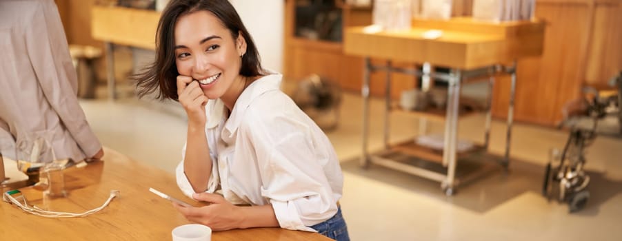 Portrait of young asian woman, sitting in cafe, holding smartphone, chatting and messaging while drinking coffee.