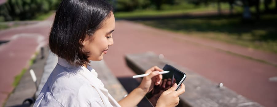 Portrait of young asian woman drawing on fresh air in park, sitting with graphic tablet and digital pen, smiling happy.