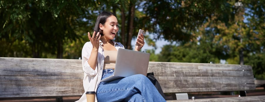 Excited young asian girl, looking at her smartphone, while sitting with laptop outdoors in sunny park.