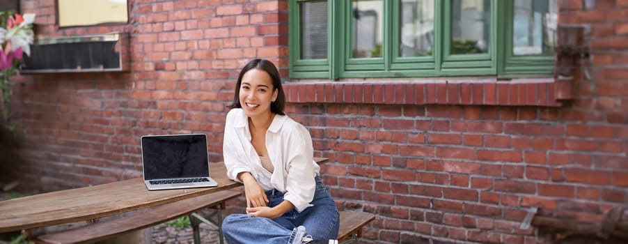 Vertical shot of good-looking asian woman with laptop, sitting in outdoor cafe, drinking coffee and smiling, laughing carefree.