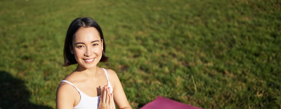 Vertical shot of young asian woman doing yoga, practice mindfulness, smiling and looking relaxed, sitting on rubber mat in park.