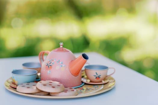 Colorful toy teapot with cups and cookies stands on a tray on a table in the garden for a tea party. High quality photo