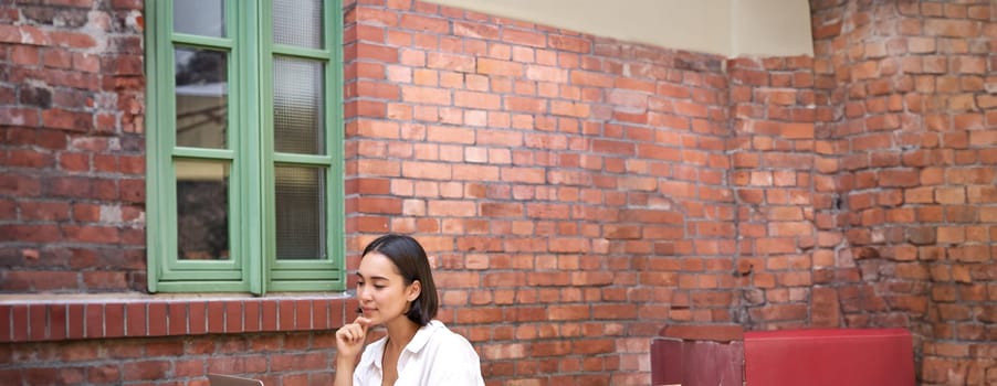 Portrait of stylish young woman, brunette girl with laptop, sitting outdoors and using computer.