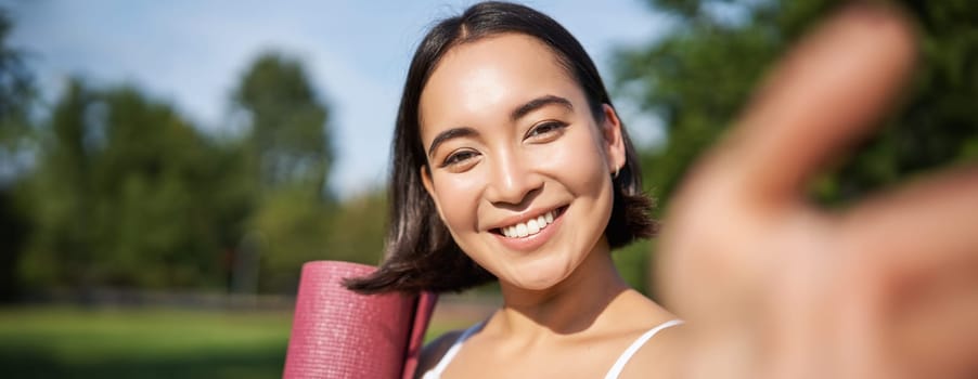 Happy asian fitness girl takes selfie with rubber yoga mat in park. Healthy young sportswoman makes photo of herself during workout.