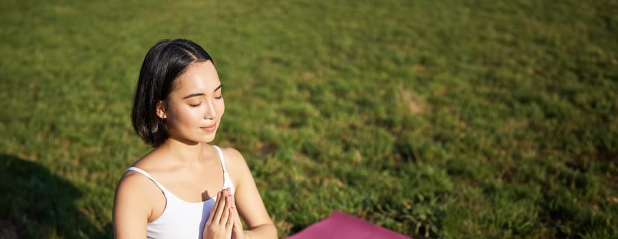 Vertical shot of young asian woman doing yoga, practice mindfulness, smiling and looking relaxed, sitting on rubber mat in park.