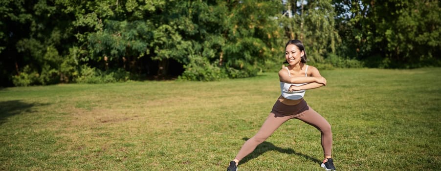 Young asian fitness girl stretching, workout in park, showing exercises, doing squats.