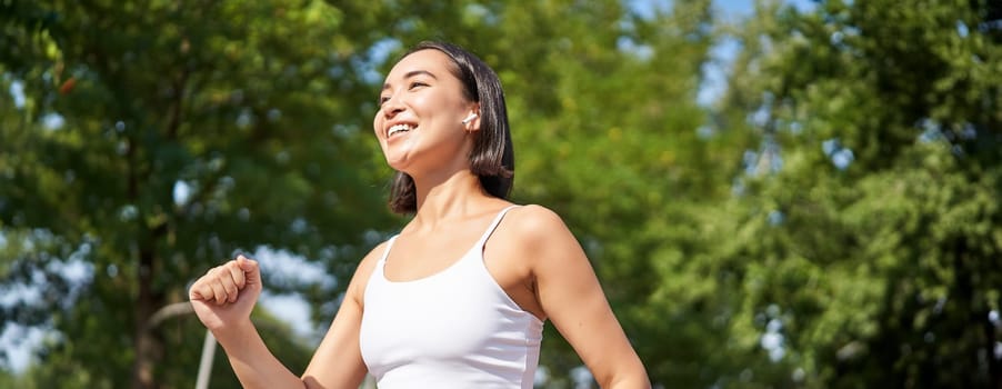Wellbeing. Smiling asian fitness girl, runner in park, smiling and running, doing jogging workout outdoors on streets.