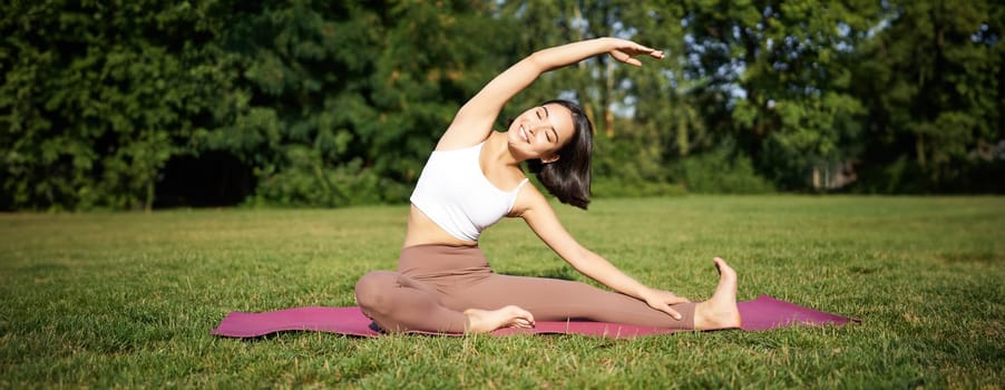 Young woman does yoga on lawn in park, stretching on fitness mat, wellbeing concept.