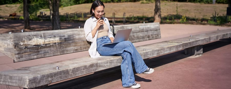 Smiling korean girl sitting in park with laptop, drinking takeaway coffee, enjoying sunny day, working or studying outdoors.