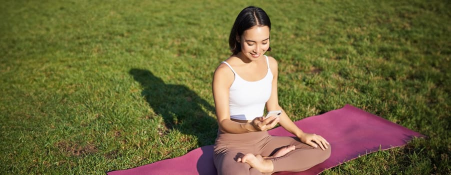 Asian girl follows yoga training app, looking at smartphone, meditating and doing exercises on fresh air in park.
