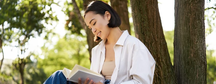 Beautiful smiling girl reading book in park on sunny day, sitting near tree on lawn and relaxing outdoors.