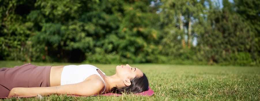 Young fitness girl lying on sport mat on lawn, breathing and meditating in park in sportswear.