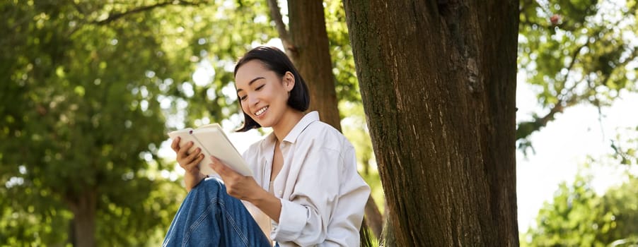 Beautiful young asian girl, student sits in park under tree and reading book, smiling, enjoying warm summer day outdoors.