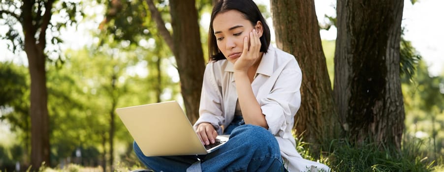 Portrait of asian woman sitting on grass near tree, using laptop, working, doing homework remotely on sunny summer day.