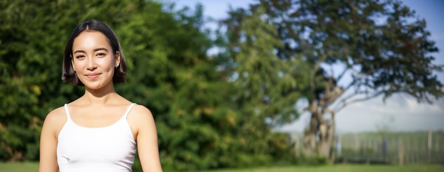 Vertical shot of young asian woman sitting on fitness mat and meditating, doing calming and relaxing exercises in park, showing zen sign.