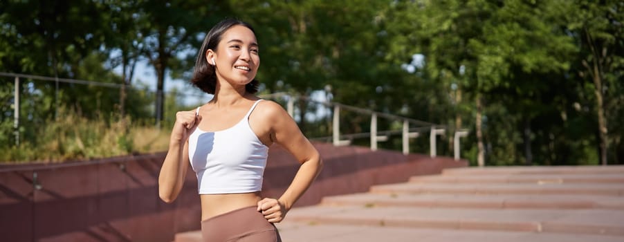 Wellbeing and sport. Asian fitness girl runner, jogging in park, running on street in leggings, smiling happily.