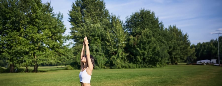 Vertical shot of smiling korean woman doing tree yoga asana, stretching on rubber mat in park, exercising.