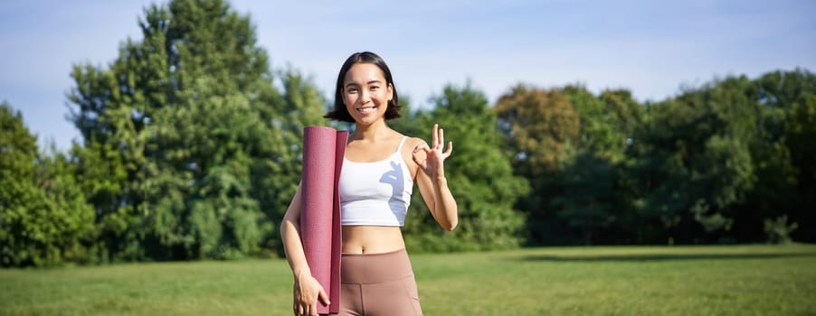 Excited young woman standing with sports mat, yoga clothes, shows okay sign, workout in park, wellbeing training session outdoors.