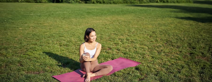 Woman in park, watching yoga video on smartphone, meditating on fresh air, sitting on rubber mat.