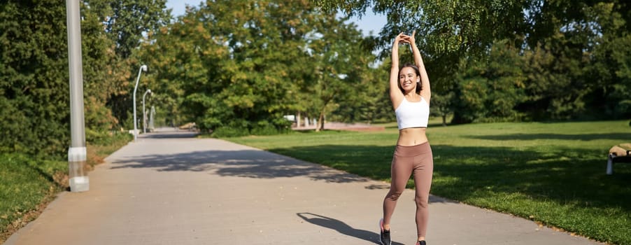 Smiling Asian girl stretching after good workout in park, listening music in wireless headphones, jogging outdoors.