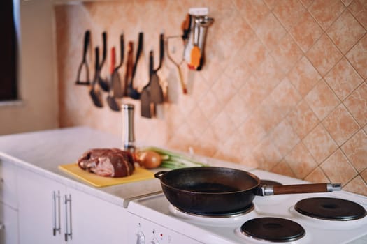 Frying pan stands on the stove against the background of a raw tenderloin and vegetables on the table. High quality photo