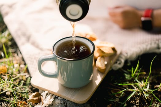 Coffee pours from a thermos into a mug standing on a wooden plank on the lawn. High quality photo